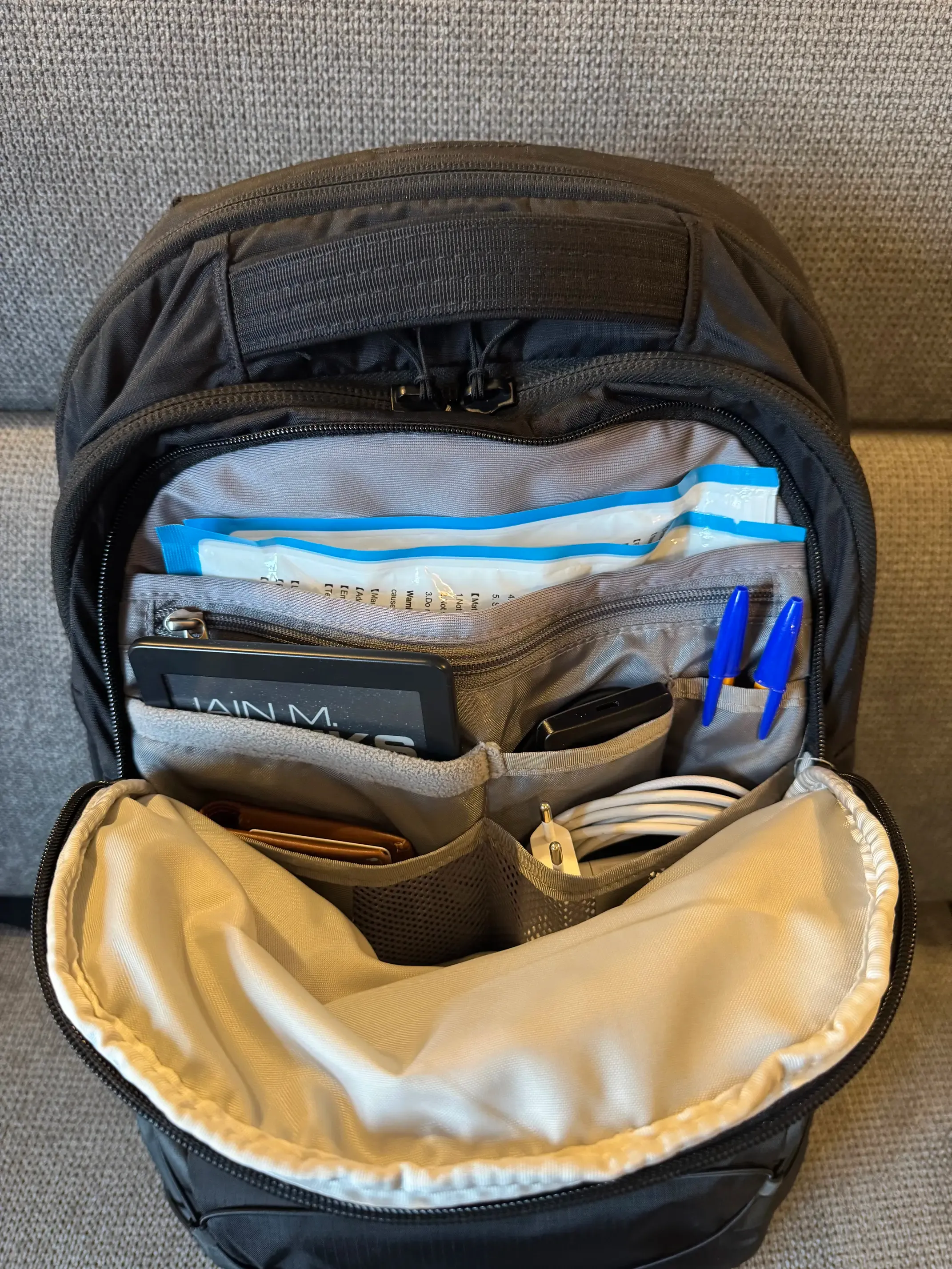 A photo of a backpack pocket with plenty of sub-pockets. There's a main pocket with FFP2 masks, a zipped pocket that takes the whole width, a medium pocket that fitsp perfectly a Kobo Clara 2E e-reader, a small pocket for a small external drive, another small pocket for two pens, and finally two medium pockets for a MacBook Pro charger and a wallet.
